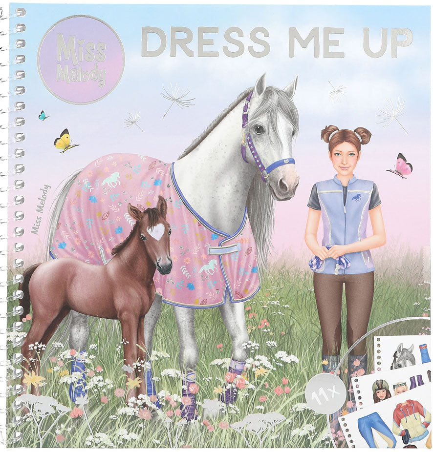 A spiral-bound children's activity book titled 'Dress Me Up' by Miss Melody, featuring an illustration of a white horse with a pink patterned blanket, a brown foal, and a girl in a blue uniform in a flower field.
