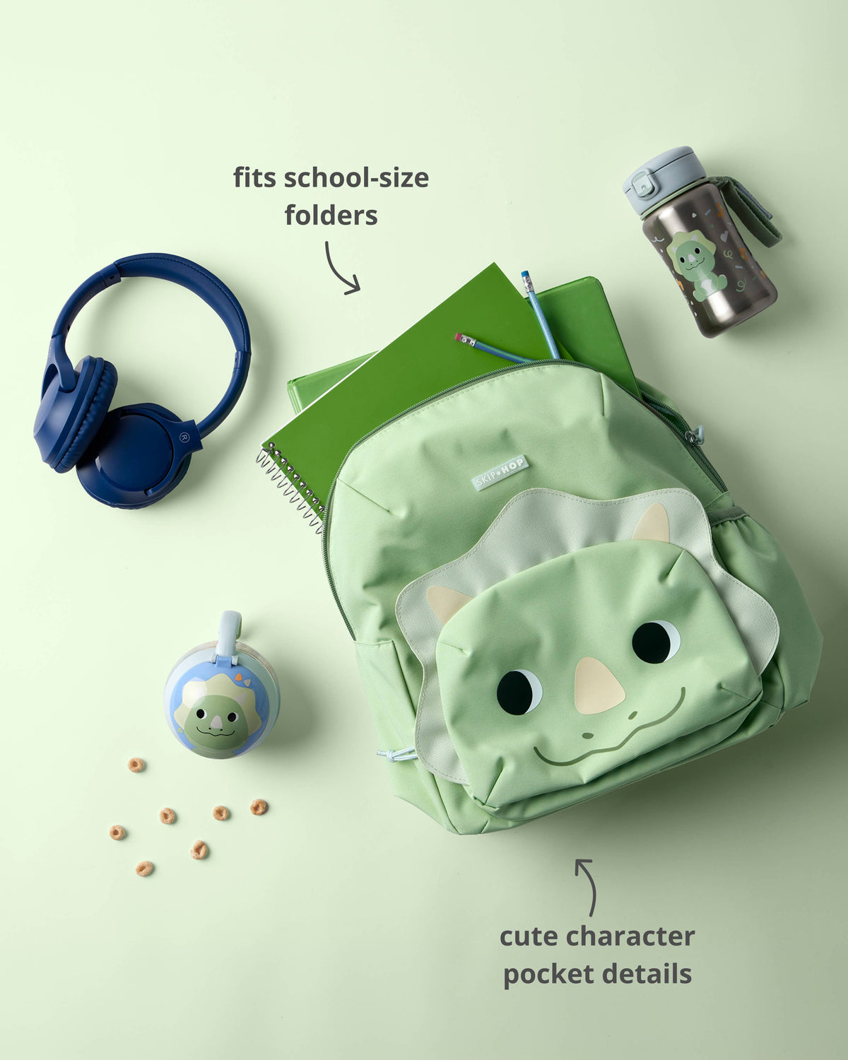 A light green dinosaur-themed backpack with a cute character face pocket is styled in a flat lay with school accessories on a pale green background.