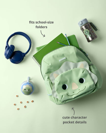 A light green dinosaur-themed backpack with a cute character face pocket is styled in a flat lay with school accessories on a pale green background.